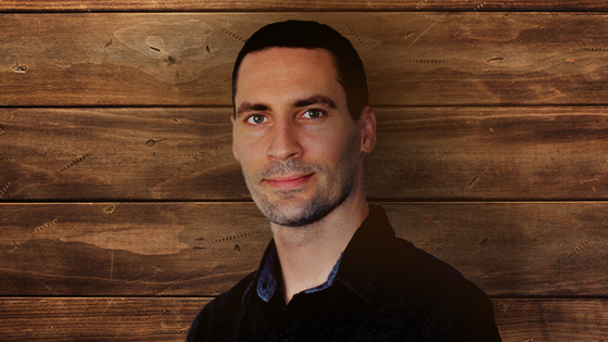 Justin Hosie standing in front of a paneled wood background. He is wearing a dark denim shirt and gently smiling.