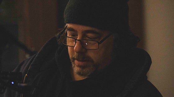 Michael Romano stands in front of a grey wall wearing a beanie, hoodie, and glasses with a pencil tucked behind his ear.