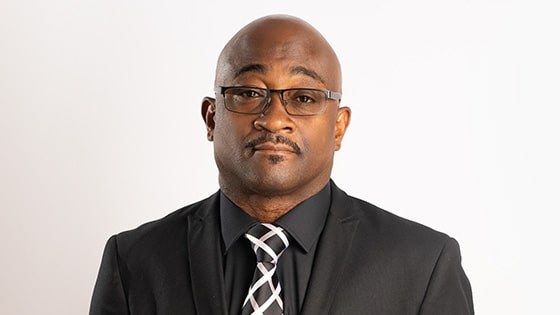 A headshot of Christopher Rapley wearing glasses and an all-black suit with a black-and-white tie and pocket square.