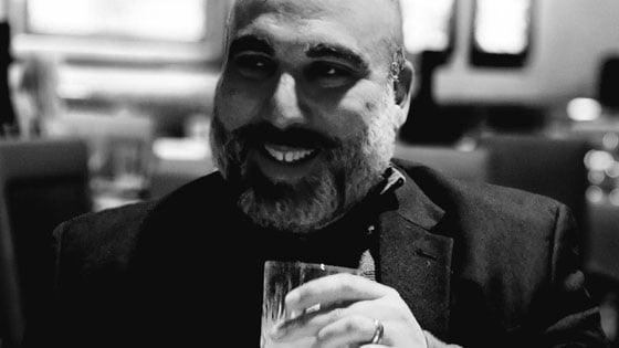 Omar Ortiz smiles as he holds a glass in his hand. He is wearing a dark jacket.
