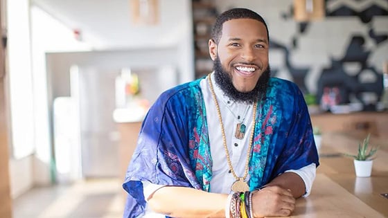 Rico Robinson stands in a café. He is wearing a blue-patterned wrap over a white button-up shirt. He is smiling.