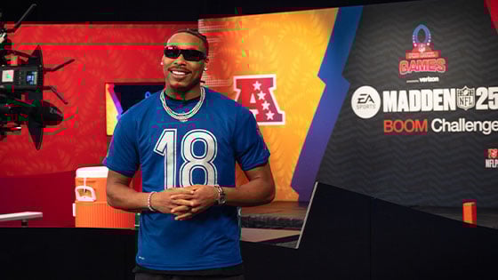 Minnesota Vikings wide receiver Justin Jefferson smiles while wearing a blue NFL-branded t-shirt with the number '18' in a studio set for the Madden NFL 25 Boom Challenge.