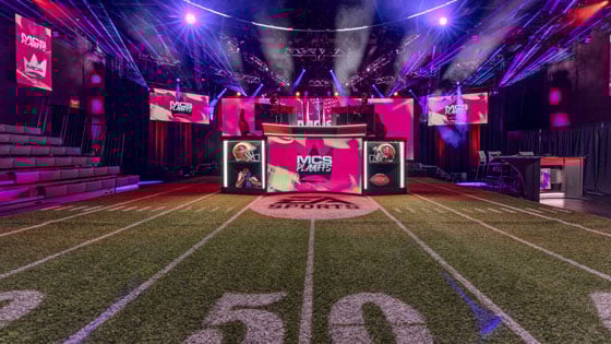 A vibrant esports stage with a football-themed setup, featuring "MCS Playoffs" branding, LED screens, and dynamic lighting.