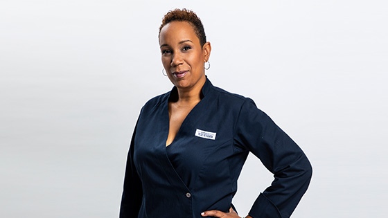 Grad Elle Simone Scott standing and smiling against a white backdrop while wearing a blue double-breasted chef's jacket with an America's Test Kitchen patch on the breast.