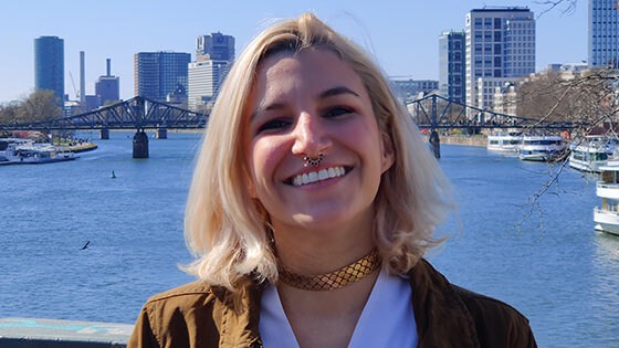 Grad Angela Decastro, a woman with shoulder-length blonde hair wearing a brown jacket and white top, smiles while standing on a bridge overlooking a river, in the distance behind her is a bridge and cityscape.