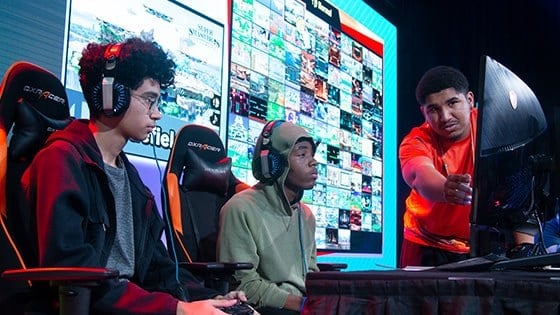 Two Florida high school students seated on stage in front of a gaming monitor with a Full Sail Armada athlete helping them to their right.