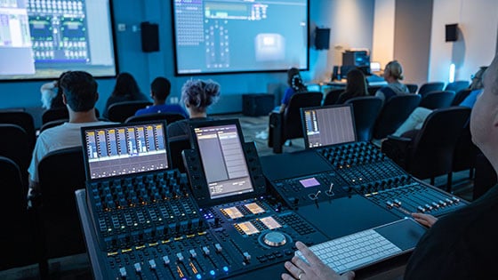 A person is seen operating a professional audio mixing console in a classroom, with students seated and screens displaying sound software.