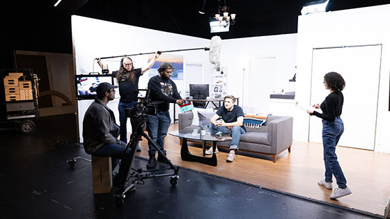 A group of students work together in a living room set on one of Full Sail’s soundstages. They are using a camera and holding microphones and a script.