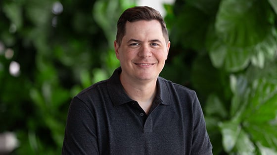 A man with short brown hair is wearing a dark grey polo while smiling against a lush green backdrop.