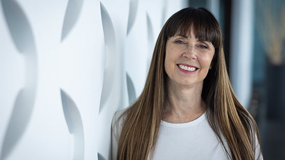 A woman with long brown hair and bangs smiling while standing against a geometric white backdrop.