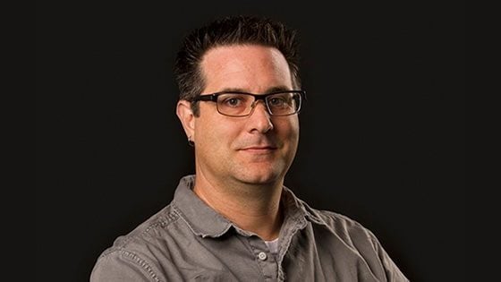 A man with short brown hair smiling at the camera while wearing black frame glasses and a gray button down against a black backdrop.