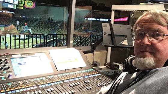 A man seated at an audio console overlooking a basketball court in a live arena.