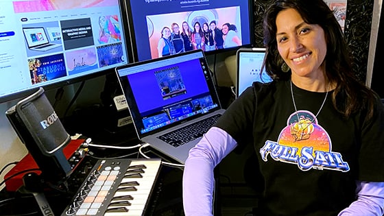 Instructor Milena Jackson, a woman with shoulder-length dark hair, is seated at her desk while wearing a Full Sail University graphic tee.