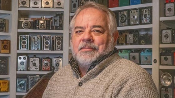 Randy sits in front of a wall covered by shelves of vintage cameras. He is wearing a light brown sweater.