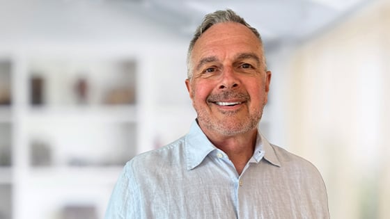 A person wearing a light blue button down while smiling against a white out-of-focus backdrop.