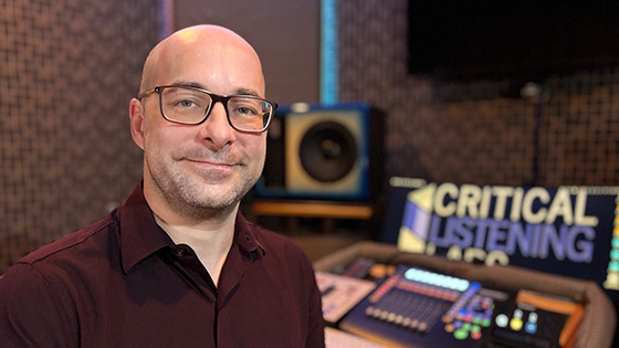 Aaron Gandia sits in a recording studio. He is smiling and wearing glasses and a burgundy shirt. A soundboard is behind him.
