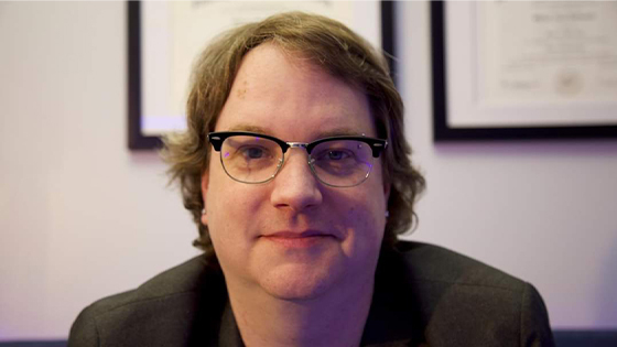 Up close headshot of a man wearing glasses and a black shirt against a wall with framed degrees.