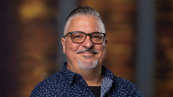 Alex Ginzo smiles at the camera. He is wearing a blue and white collared shirt and glasses, and he has gray hair and a short gray beard.