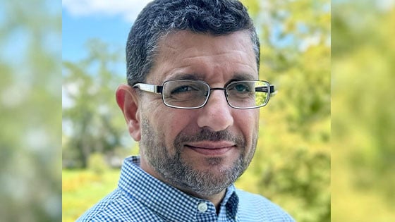 A man with short curly hair, glasses, and a trimmed beard smiles slightly while standing outdoors, the background is out of focus.