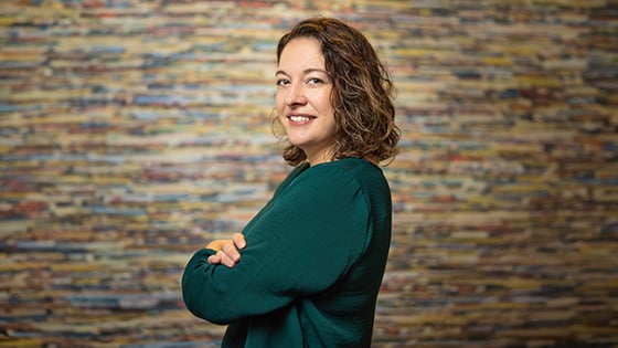 Aron Gelineau stands with her arms crossed and smiles at the camera. She has brown, curly hair and is wearing a teal sweater.