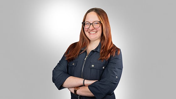 A smiling person with long red hair, glasses, and a blue zip-up jacket, is standing with their arms crossed against a light gradient background.