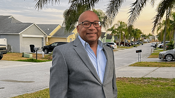 Chris stands outside wearing a grey blazer, blue shirt, and black-rimmed glasses. He is smiling.