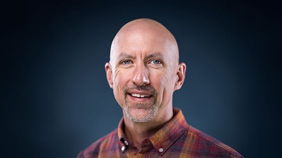 Corbett Compel smiles for the camera in front of a blue background. He is wearing a plaid shirt.