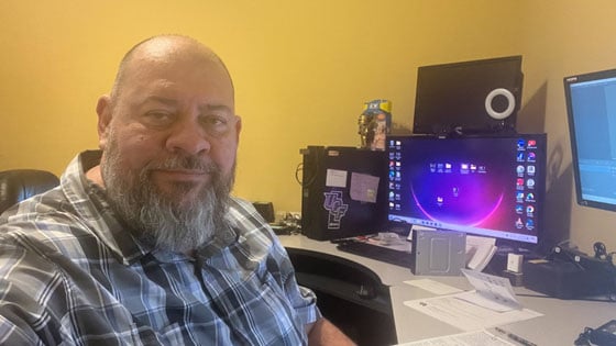 Dan Walker sits in his home office. He has a beard and is wearing a plaid shirt. A desk with several desktop monitors is behind him.