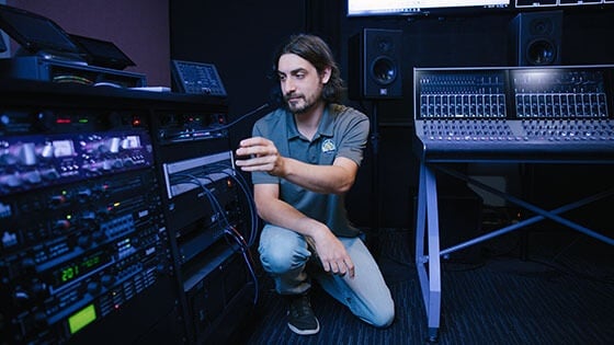 Dave Harris crouches next to broadcast equipment, including mixing consoles and a communications system.