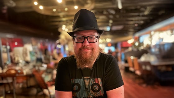 David Dennison sits at a table with his arms crossed. He has glasses and a beard and is wearing a black t-shirt and black fedora.