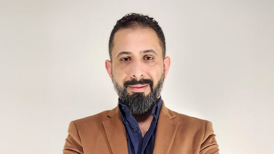 Dr. Ahmed Al Zaidy stands with his arms crossed and smiles at the camera. He is wearing a brown blazer over a blue button-up shirt.