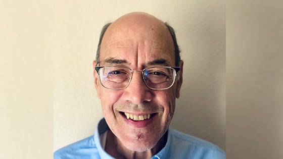 Dr. Carstens stands in front of a white wall wearing a blue shirt and glasses. He is smiling.