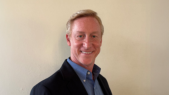 Greg Downey smiles at the camera in front of a light background. He is wearing a blue shirt and a dark blazer.