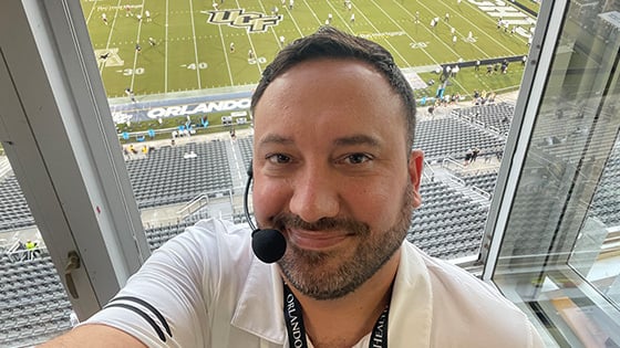 Jeff Sharon wears a headset microphone and smiles at the camera. He is in an announcer’s booth with the UCF football stadium below him.