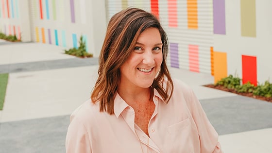 Jenn Miller sits in front of a colorful outdoor wall. She is smiling and wearing a pink shirt and a gold necklace.