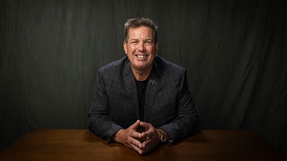 Joe Gornik sits at a wooden table and smiles at the camera. He is wearing a gray plaid jacket with a Full Sail employee pin on the lapel.