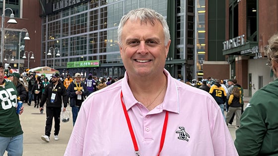 A man with short white hair and a pink polo shirt is seen smiling while standing in front of Lambeau Field in Green Bay, Wisconsin.