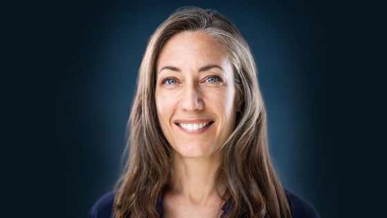 Kelly sits in front of a blue background wearing a blue blouse. She has blue eyes and is smiling.