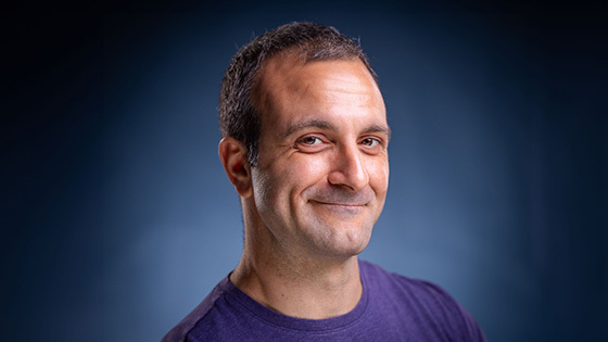 Michael Corinella smiles at the camera in front of a blue background. He is wearing a purple shirt.
