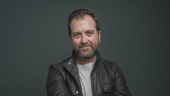 A man in a dark brown leather coat and light t-shirt appears from the waist up, smiling against a grey backdrop.