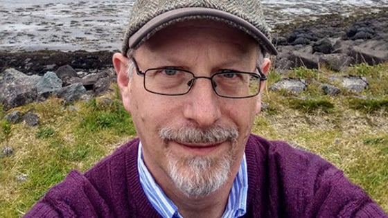 Peter Gordon stands outdoors in front of a river. He is wearing a purple sweater, a newsboy cap, and glasses.