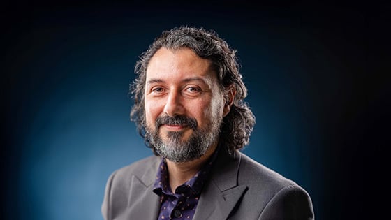 Plinio Pinto smiles at the camera. He has curly brown hair and a beard streaked with gray, and he wears a gray blazer.