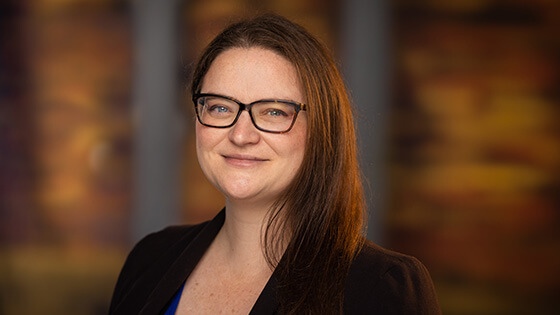 Rebecca Leis has brown hair and is wearing tortoiseshell glasses and a black blazer. She is smiling.