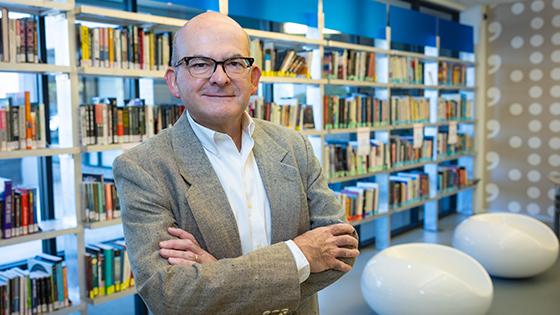 Rolando stands in a library with his arms crossed. He is smiling and wearing a suit.