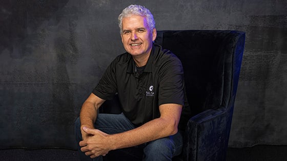 Roy Papp sits in a blue velvet chair in a photography studio. He is smiling and wearing jeans and a black polo shirt with a Full Sail logo.