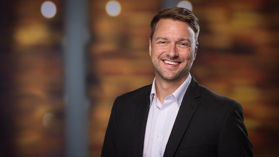 A smiling man in a black blazer and light dress shirt stands in front of a warmly lit, blurred background.