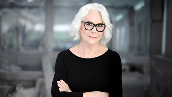 A woman with short white hair and glasses, wearing a black top, smiles confidently with arms crossed against a blurred background.