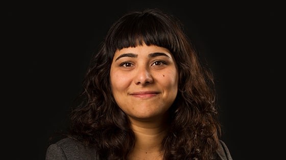 Suzy Johnson wears a gray blazer and a silver necklace and sits against a black background. She is smiling.