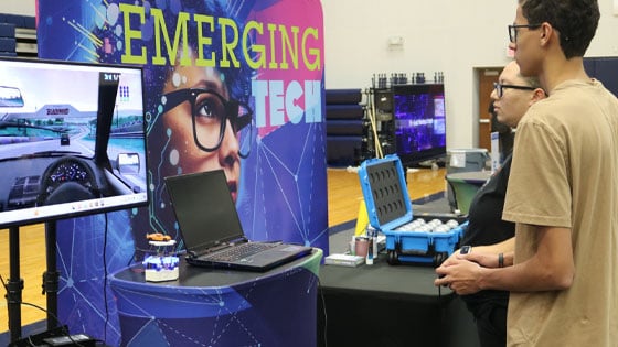 Two people at TechFest explore a driving simulator next to an ‘Emerging Tech’ display with gadgets and a laptop on the table.