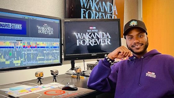 Alain sits at a desk with multiple monitors that read “Wakanda Forever” and “Marvel Studios.” He is wearing a purple hoodie.